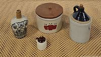 Group photo showing all four items on woven mat: two stoneware crock jugs, apple painted crock with lid, and antique whiskey jug.