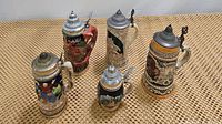 Photo showing set of five German beer steins in a grouping on a woven mat surface. Each has a pewter hinged lid and varied colorful designs including a fox handle on one stein.