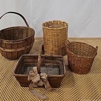 Four decorative baskets displayed on a textured surface: wooden apple basket, cylindrical wicker waste basket, rectangular dark wicker handled basket with bow, and round wicker basket with handles.