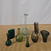 Full view of all eight vases arranged on woven surface showing variety of shapes and shades of green glass.