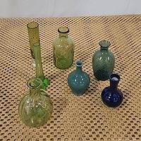 Seven assorted glass vases in green, aqua blue and cobalt blue colors arranged on woven mat.