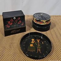 Three items on a woven mat: black folk art painted tin with handles, round black folk art painted tray with a couple image, and black antique cake box with orange floral band and glass knob lid.