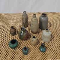 View of 10 ceramic jugs and vases arranged on a woven surface showing various shapes, colors, and sizes.