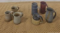 Nine mugs arranged on woven surface showing all mugs together.