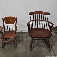 Pair of two wooden spindle back rocking chairs on concrete floor, small child's and larger Windsor style captain's chair side by side.