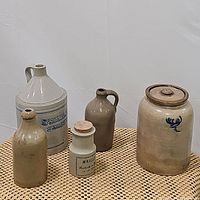 Five stoneware pieces arranged on woven mat: Shepard jug, crock with lid, two jugs, bottle and Maille jar