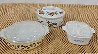 Photo showing all three vintage casserole dishes on a woven surface: Corningware oval Ivy pattern casserole, Evesham round fruit pattern casserole, and Corningware blue cornflower square casserole.