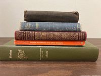Five old books stacked horizontally showing visible titles and wear.