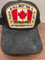 Front view of DSquared2 black baseball hat. Patch shows Canadian flag and bold text including profanity, branding, and 'One Voice Speaks For Many'. Hat fabric appears dusty and worn.