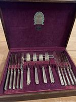 Full view of the flatware set inside the wooden box with burgundy fabric lining. Shows the arrangement of knives and forks.