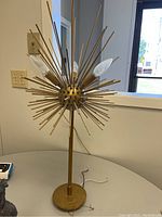 Full view of the gold starburst table lamp showing the central spherical metal core with numerous gold rods radiating out and three candle bulbs installed in socket holders extending from the center. Lamp is standing on a round base on a grey table in a room with a window and light switch visible in the background.