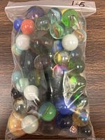 Plastic bag containing multiple vintage glass marbles of various colors and patterns, including green, blue, white, black, and swirled designs; marbles vary in size and transparency.