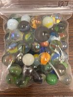 Clear plastic bag containing approximately 40 assorted vintage glass marbles in various colors and patterns, including green, white, black, yellow, orange, and translucent ones with swirls and speckles.