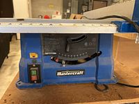 Close-up of the base of the blue Mastercraft table saw showing the power switch, brand logo, and adjustable blade angle indicator.