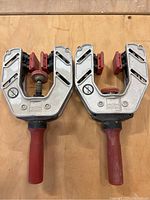 Top view of both Bessey one-handed edge clamps placed side by side on a wooden surface showing their metal bodies, red handles, and protective red jaw pads.