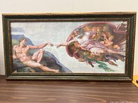 Framed reproduction print of 'The Creation of Adam' by Michelangelo, showing the entire artwork within a decorative frame placed on a wooden table surface.