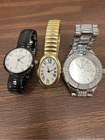 Three Geneva watches side by side on wood surface, showcasing different styles and colors: black face watch, gold-tone oval watch, silver-tone crystal watch.