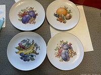 Four plates showing different fruit designs including plums, grapes, bananas, oranges, and peaches.