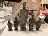 Five terracotta warrior figurines arranged in a row including one larger central figure and four smaller ones, placed on a table next to a water bottle for size reference.