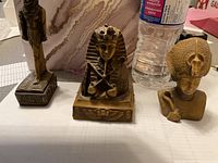 Three Egyptian-themed statuettes with detailed features and varying sizes placed on a white surface next to a water bottle for scale.