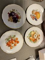 Top view showing 4 fruit-themed plates with gold rim and scalloped edges on carpet