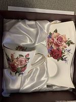 Tea cup and rectangular saucer with floral rose design and gold trim, displayed on white satin fabric inside the box.