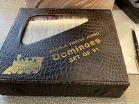 Photo showing textured black box with gold embossed text 'Double Twelve Jumbo Dominoes Set of 91' and a see-through window displaying contents inside.
