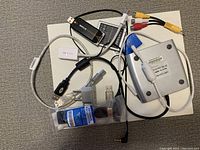 Top view of assorted electronic items with cables, adapters, and a small gray device with antenna placed on a white surface.