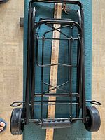 Folded hand trolley laid flat showing metal frame and wheels beside a yardstick for size reference.