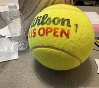 Oversized tennis ball with Wilson US Open branding placed next to a water bottle for scale, showing it is much larger than a standard ball.
