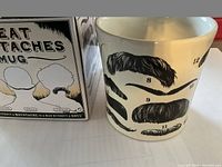 Photo showing the side of the white mug with six different black mustache designs, each numbered, accompanied by the original box to the left.
