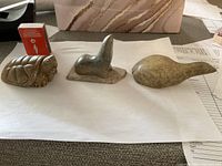 Overview of three small stone animal carvings including a turtle, a seal and a bird placed on white paper with a matchbox for scale.
