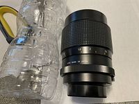 Side view of black camera lens showing aperture ring and 'Lens Made in Japan' engraving, placed next to a plastic water bottle for scale.