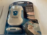 Front view of ManiPedi Elite nail file and shaper kit in sealed packaging. Shows white and blue device with finger holder and pen-shaped buffer.