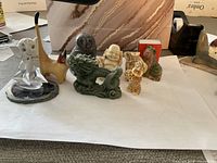 All seven small figurines displayed on a white surface with background items including a pink bag, tape dispenser, and papers. Figurines vary in size, color, and material.