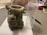 Side view of the stone carved vase showing carved floral decoration and four rounded feet at base, alongside a water bottle for size reference.