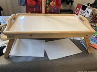 Wood bed tray table showing angled side view with fold out legs, natural wood finish frame and white surface.