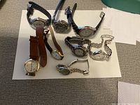 Wide shot showing nine wrist watches laid on white paper on carpet. Various metal, leather, fabric, and rubber bands observable. Watches mostly round face, various sizes.