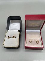 Two pairs of 14k gold stud earrings displayed inside two different earring boxes; one pair rectangular box-shaped studs and one pair delicate open heart-shaped studs.