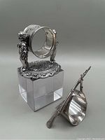 Napkin ring with two cherubs holding the ring, sitting on an ornate circular base, placed on a clear cube stand beside the rifle napkin ring.