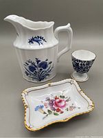 All three Royal Crown Derby items: Festival Blue creamer jug, Derby Posies trinket dish, antique chinoiserie egg cup against neutral background.