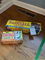Photo showing the two boxed sets of sports cards on a wooden floor: a large yellow box for 1986 Topps Baseball Cards and a smaller yellow box for 1988 Topps Football Cards.