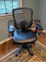 Front view of black office chair with mesh backrest, padded seat and armrests, positioned near a window.