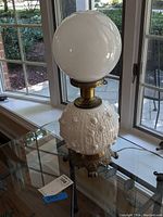 Side view of 20-inch Fenton milk glass lamp with large white milk glass globe and ornate dark metal base on glass table near window.