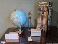 Photo showing the globe, Galileo thermometer, storm glass, and boxed hanging thermometer displayed on a glass tabletop.