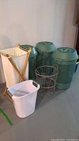 Photo showing three green lidded laundry hampers, one wooden frame fabric hamper, one white plastic basket with handles, and one metal wire basket frame