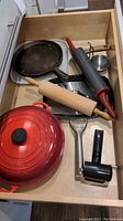 Drawer with cast iron cook pot, rolling pins, a round cast iron griddle, a metal peeler, a black rolling cutter, and a small rectangular metal tray.