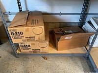 Two unopened and one partially opened cardboard boxes on shelving. Two boxes labeled Libbey 8418 Grande with 1 dozen stemmed wine glasses each; one box partially filled with plastic bottles and screw caps.