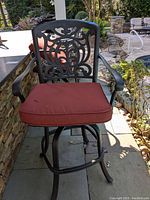 Close up of one patio bar stool showing metal decorative backrest, armrests, and red seat cushion with tie strings.