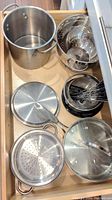 Top-down photo of stainless steel cookware including large pot, colander, smaller pot, lids, and perforated insert inside kitchen drawer.
